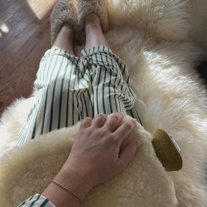 May include: A pair of cream-coloured fuzzy shearling slippers and white trousers featuring vertical dark green stripes. The slippers have a thick, textured pile, and the trousers are made of a lightweight fabric with a crisp, clean pattern.