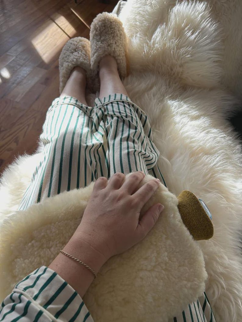 May include: A pair of cream-coloured fuzzy shearling slippers and white trousers featuring vertical dark green stripes. The slippers have a thick, textured pile, and the trousers are made of a lightweight fabric with a crisp, clean pattern.