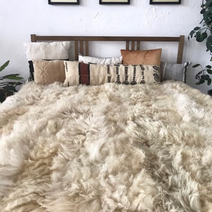 Pode incluir: Um tapete de pele de carneiro branco com textura felpuda, colocado em uma cama com cabeceira de madeira e v&aacute;rios travesseiros estampados.
