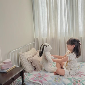 May include: A plush stuffed rabbit toy with cream-colored fur and dark brown ears. The rabbit has a soft, textured appearance and is held by a child sitting on a bed with a white metal frame, a colorful patchwork quilt, and decorative pillows.