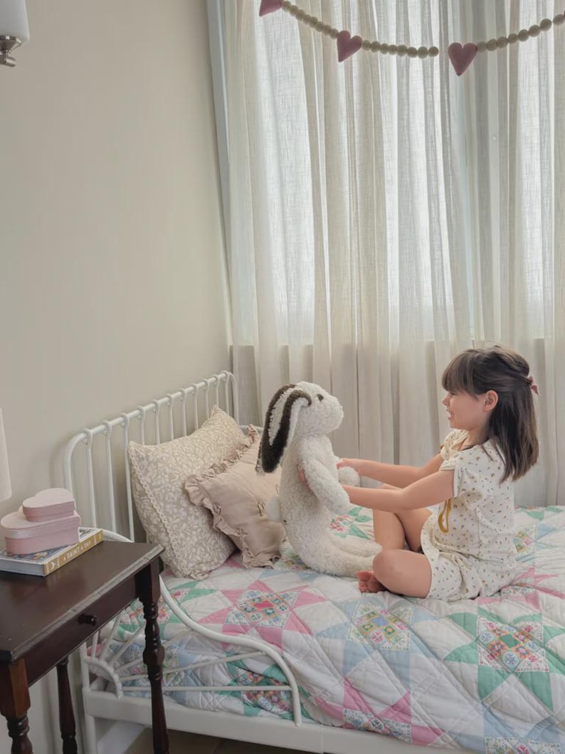 May include: A plush stuffed rabbit toy with cream-colored fur and dark brown ears. The rabbit has a soft, textured appearance and is held by a child sitting on a bed with a white metal frame, a colorful patchwork quilt, and decorative pillows.
