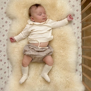 May include: A baby sleeping on a white sheepskin rug with a white ruffled shirt, brown shorts, and white socks.