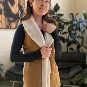 May include: A brown faux fur vest with a white faux fur collar and lining. The vest has a zipper closure and is worn over a black long-sleeved shirt and blue jeans.
