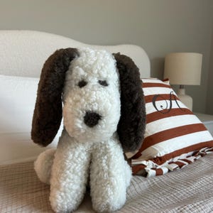 May include: A plush dog toy with white fur and brown ears, nose, and eye details. The stuffed animal is sitting on a bed with a striped pillow and a lamp in the background. The dog toy is approximately 10 inches tall.