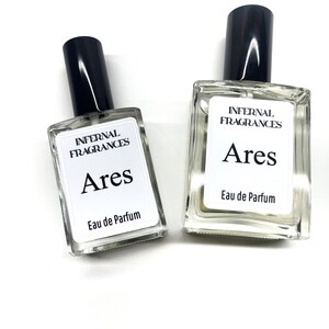 Puede incluir: Dos botellas de perfume de vidrio transparente con tapas negras y etiquetas blancas. Las etiquetas dicen "Infernal Fragrances Ares Eau de Parfum".