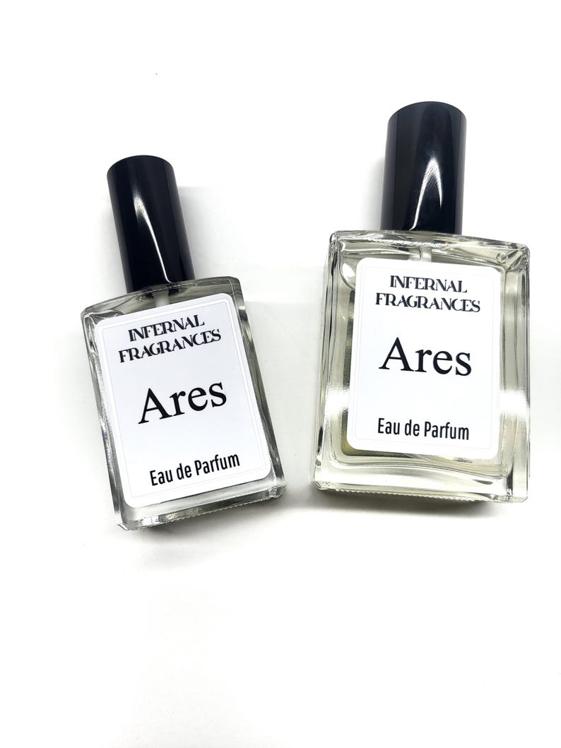 Puede incluir: Dos botellas de perfume de vidrio transparente con tapas negras y etiquetas blancas. Las etiquetas dicen "Infernal Fragrances Ares Eau de Parfum".