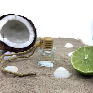 May include: A clear glass bottle with a wooden cap and a gold cord hanging from it. The bottle is sitting on a bed of sand with a half coconut, a lime wedge, and seashells.
