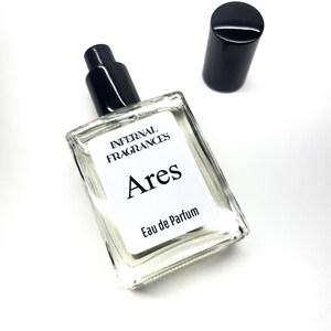 Puede incluir: Una botella de perfume de vidrio transparente con una tapa negra y una etiqueta. La etiqueta dice "Infernal Fragrances Ares Eau de Parfum".
