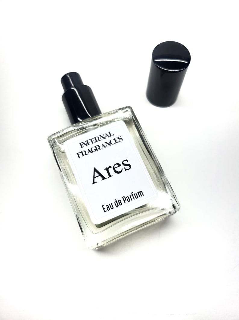 Puede incluir: Una botella de perfume de vidrio transparente con una tapa negra y una etiqueta. La etiqueta dice "Infernal Fragrances Ares Eau de Parfum".