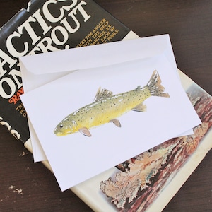 Puede incluir: Una tarjeta de felicitación blanca con una ilustración de acuarela de una trucha. La tarjeta está colocada encima de un libro con el título "Tactics on Trout" y un sobre blanco. Tema rústico y de naturaleza.