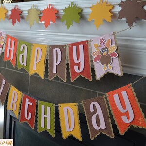 May include: A festive banner with the words "Happy Birthday" in pink letters on brown, orange, yellow, and green pennants. A turkey with a pink bow is on one of the pennants.