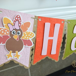 May include: A Thanksgiving banner with the word "Happy" in pink letters on a brown, orange, and green background. The banner features a cute cartoon turkey with a pink bow.