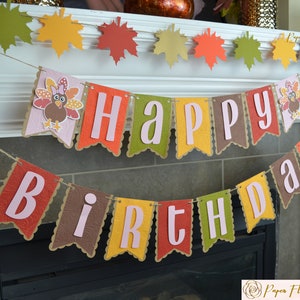 May include: A festive banner with the words "Happy Birthday" in pink letters on a brown, green, yellow, and red background. The banner features a turkey design with a pink bow and feathers.
