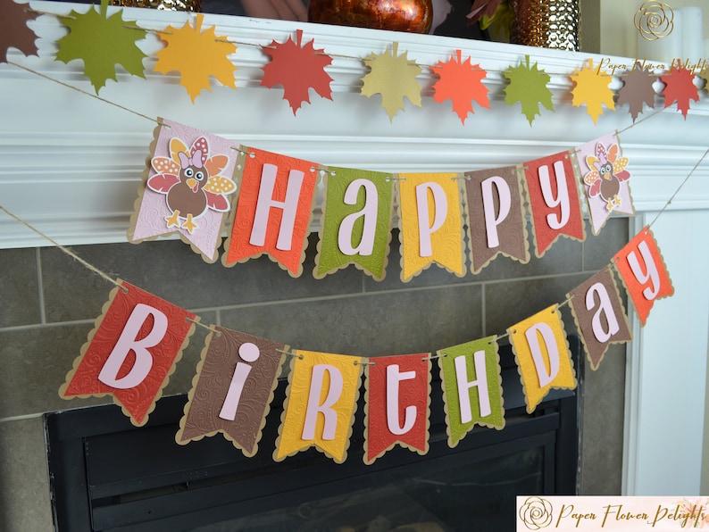 May include: A festive banner with the words "Happy Birthday" in pink letters on a brown, green, yellow, and red background. The banner features a turkey design with a pink bow and feathers.
