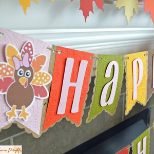 May include: A pink, orange, and brown paper banner with the word "HAPPY" in pink letters. The banner is decorated with a brown turkey with pink feathers and a pink bow.