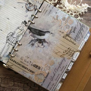 May include: Open journal with vintage-style pages. The pages feature illustrations of a bird and a woman, along with handwritten text and decorative lace. The journal is bound with metal rings and has monthly tabs.