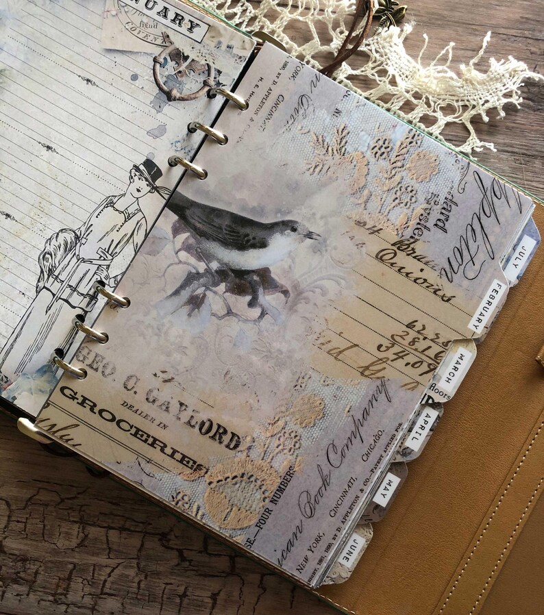 May include: Open journal with vintage-style pages. The pages feature illustrations of a bird and a woman, along with handwritten text and decorative lace. The journal is bound with metal rings and has monthly tabs.