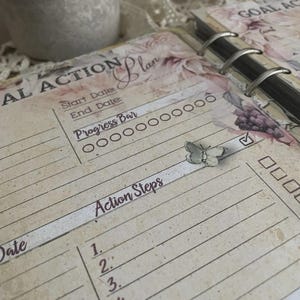 May include: A vintage-style planner page with floral accents and the words "GOAL ACTION Plan" and "Action Steps." A small butterfly embellishment sits on the page. The page includes a progress bar and check boxes.