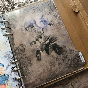 May include: Open leather binder with a floral design insert. The insert features a vintage-style illustration of blue roses and green leaves on a textured background. A tab labeled "DECEMBER" is visible.