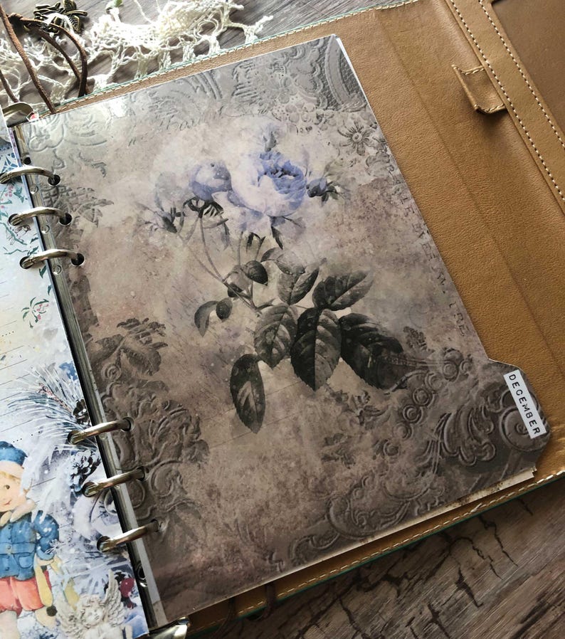 May include: Open leather binder with a floral design insert. The insert features a vintage-style illustration of blue roses and green leaves on a textured background. A tab labeled "DECEMBER" is visible.