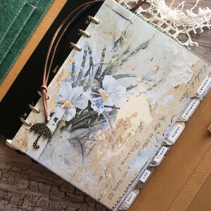 May include: A vintage-style planner with a floral design and monthly tabs. The cover features a watercolor painting of white flowers with blue accents and handwritten script. A brown leather cord with an umbrella charm is attached. The planner is open to the January page.