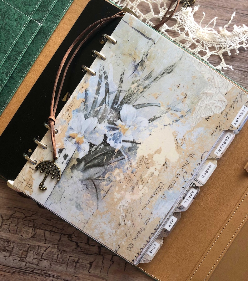 May include: A vintage-style planner with a floral design and monthly tabs. The cover features a watercolor painting of white flowers with blue accents and handwritten script. A brown leather cord with an umbrella charm is attached. The planner is open to the January page.