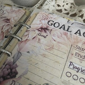 May include: Open planner pages with floral designs and the words "GOAL ACTION" and "Goal" printed on the pages. The pages have a vintage aesthetic with a cream background and pink and purple floral accents. The planner is held together with silver metal rings.