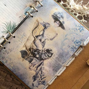 May include: A vintage-style planner with a printed illustration of a woman in a hat. The pages have a floral and script design with month tabs. The planner is bound in a brown leather cover.