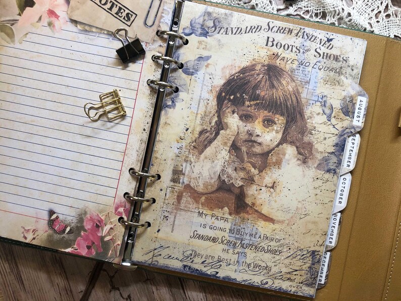 May include: Open journal with lined pages, binder rings, and a vintage-style illustration of a child. The journal features a floral design, binder clips, and monthly tabs. The illustration includes text that reads "STANDARD SCREW FASTENED BOOTS SHOES HAVE NO EQUAL."