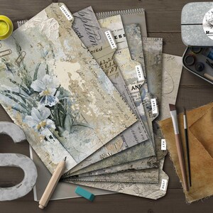 May include: An artistic still life featuring a stack of decorative papers labeled with months, a metal tin with art supplies, paint tubes, brushes, and a large letter "S". The papers have floral and script designs.