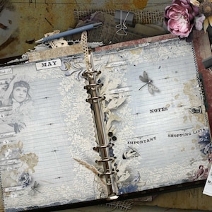 An open ring-bound planner with vintage-style graphics, including a portrait of a person, a dragonfly, and lace patterns. The pages feature the text MAY, NOTES, IMPORTANT, and SHOPPING LIST, with sections for days of the week.