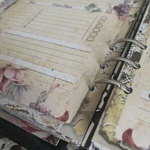 May include: Open planner pages with lined sections, checkboxes, and floral accents. The pages have a vintage aesthetic with a distressed, aged look. Silver binder rings hold the pages together. Floral and lace embellishments add a decorative touch.