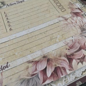 May include: A vintage-style journal page with the words "Action Steps" and a checklist. The page has a floral design in shades of pink and cream, with a distressed, aged appearance. The journal page is lined and has a decorative lace trim.
