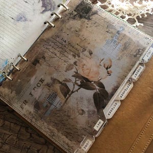 May include: A vintage-style planner with a floral design and month tabs. The pages feature a faded, antique aesthetic with a rose illustration and text. The planner is bound with metal rings and has a leather cover.