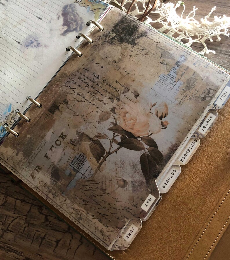 May include: A vintage-style planner with a floral design and month tabs. The pages feature a faded, antique aesthetic with a rose illustration and text. The planner is bound with metal rings and has a leather cover.