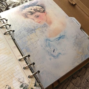 May include: Open planner with a vintage-style illustration of a woman in a blue dress. The page has a light blue and white background with the words "October" and "November" on the tabs. The planner is bound with metal rings.