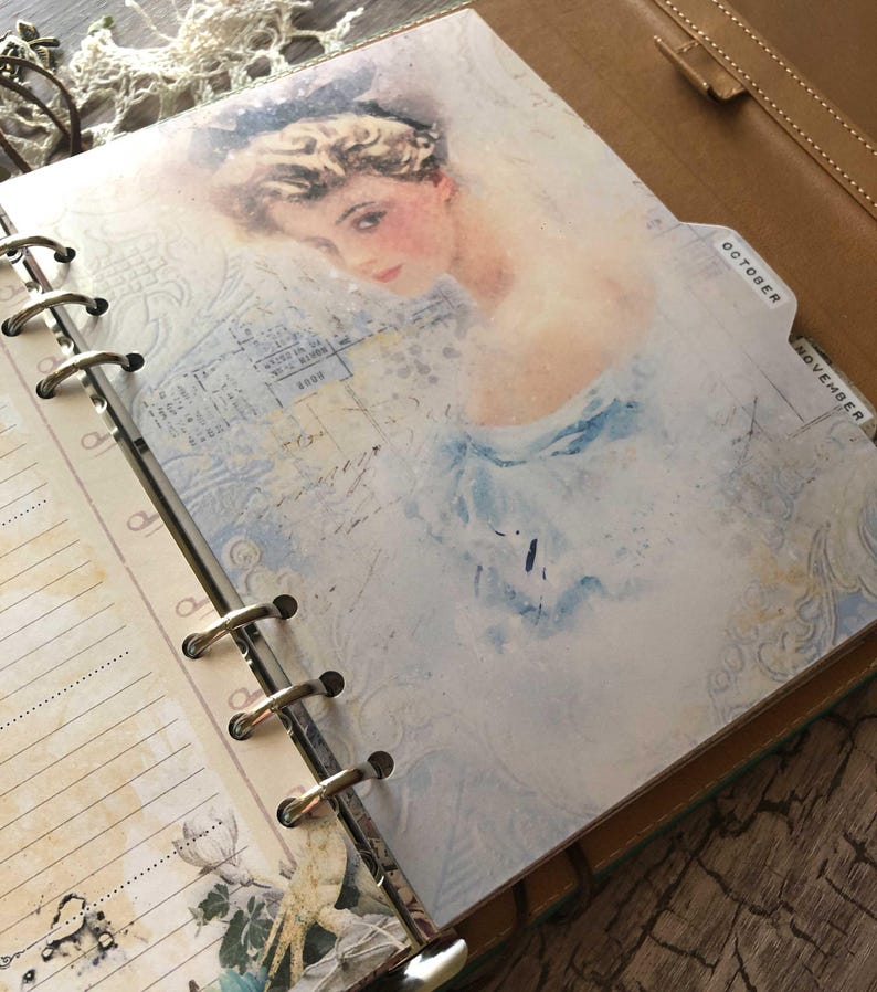 May include: Open planner with a vintage-style illustration of a woman in a blue dress. The page has a light blue and white background with the words "October" and "November" on the tabs. The planner is bound with metal rings.