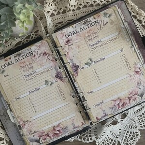 May include: Open planner with floral design pages. The pages are labeled "Goal Action Plan" with sections for start and end dates, progress bars, action steps, and notes. The planner has a ring binder and is decorated with butterfly accents.