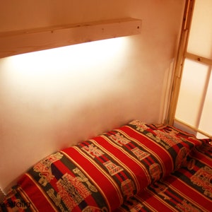 May include: A wooden shelf with a light fixture attached to it. The shelf is mounted on a wall above a bed with a red and gold patterned bedspread.