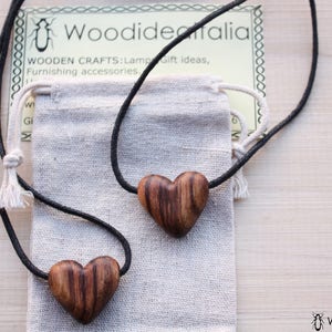Puede incluir: Dos collares de madera con forma de corazón con cordones negros y una pequeña bolsa de tela. Los collares están hechos por Woodideitalia, una empresa que fabrica artesanías de madera, lámparas, ideas para regalos y accesorios de decoración.