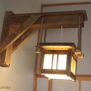 Puede incluir: Una lámpara de pared de madera con una linterna cuadrada colgando de ella. La linterna está hecha de madera y tiene un panel de vidrio blanco esmerilado. La lámpara está fijada a la pared con un soporte de madera y está suspendida por una cuerda.
