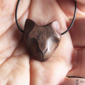 Puede incluir: Un colgante de madera con forma de cabeza de gato en un collar de cordón negro. El colgante está tallado en madera oscura y tiene una cara de gato realista.