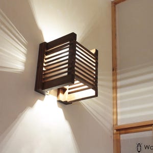 Pode incluir: Arandela de parede de madeira marrom escuro com design de ripas, projetando padrões de luz e sombra na parede. A luminária tem formato quadrado e é montada em uma parede branca. Elemento decorativo.
