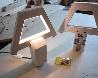 May include: Two wooden lamps with a triangular frame and a square light panel. The lamp on the left is lit and has a white cord and switch. The lamp on the right is unlit and has a visible electrical component. A small bag of screws is in the lower right corner.