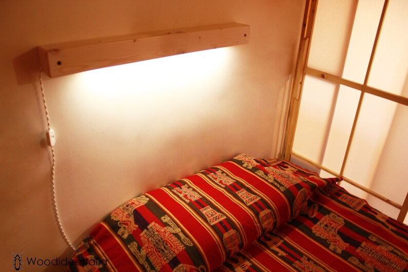 May include: A wooden shelf with a light fixture attached to it. The shelf is mounted on a wall above a bed with a red and gold patterned bedspread.