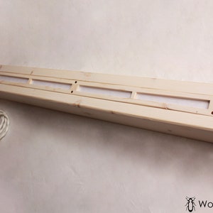 May include: A long, rectangular wooden light fixture with a white cord and switch. The fixture has three rectangular openings with a white material behind them.