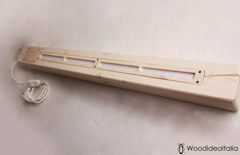 May include: A long, rectangular wooden light fixture with a white cord and switch. The fixture has three rectangular openings with a white material behind them.