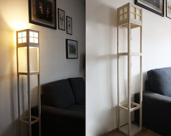 May include: A wooden floor lamp with a white shade and multiple shelves. The lamp is lit and casting a warm glow. The lamp is in a living room setting with a couch and framed artwork on the wall.