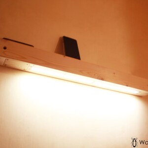 May include: A wooden shelf with a built-in light fixture. The shelf is mounted to a wall and has a small phone resting on it. The light is turned on and illuminates the shelf.