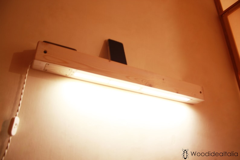 May include: A wooden shelf with a built-in light fixture. The shelf is mounted to a wall and has a small phone resting on it. The light is turned on and illuminates the shelf.